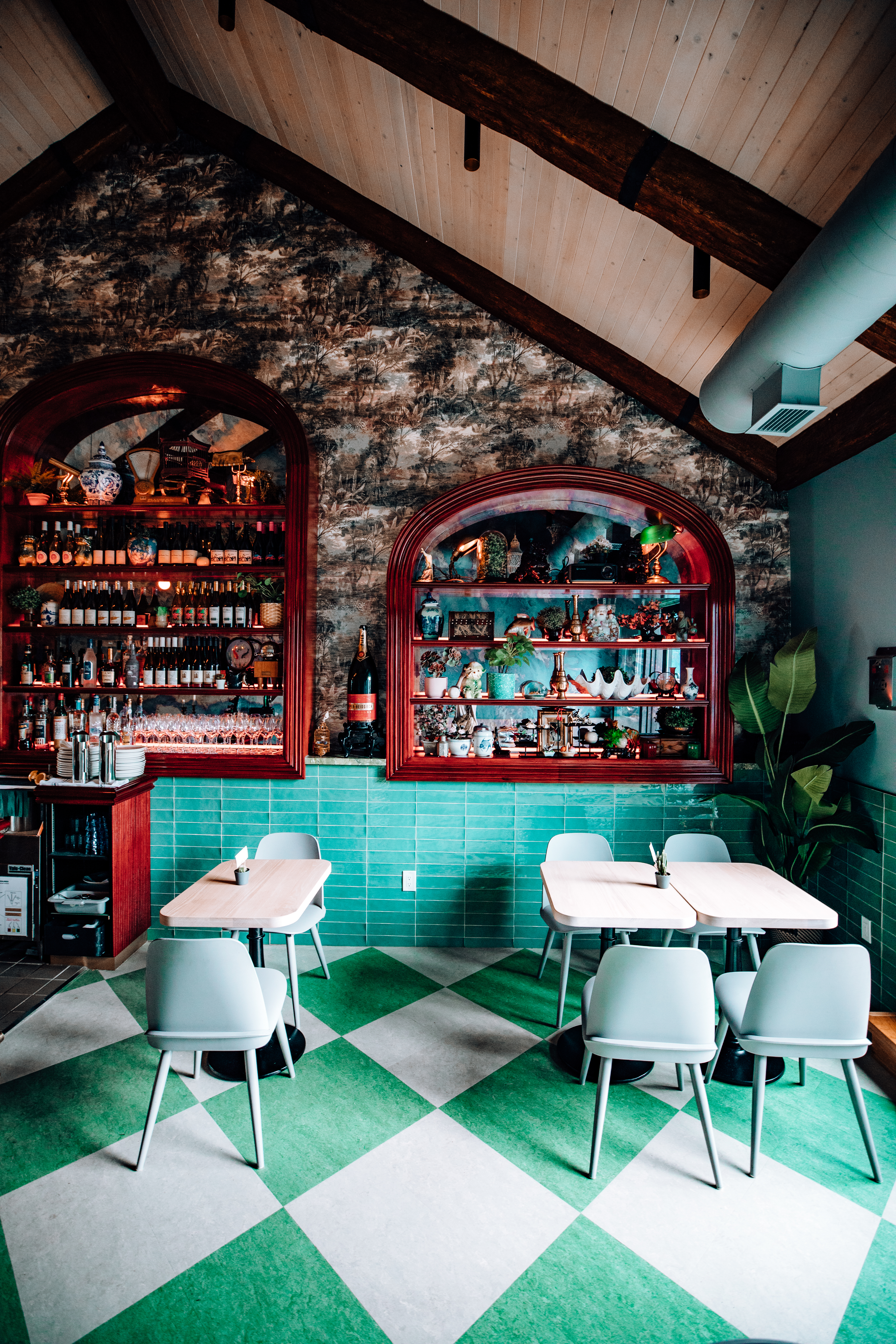 Bar Mignonette Interior. Sea green and white checker tile floors. Sea green subway tiles line 1/4 of the back wall from the bottom. The top 3/4 of the back wall has graphic wallpaper with plants drawn in thin brushstroke pattern. There arched bar shelves lined with liquor bottles.Room has vaulted ceilings.