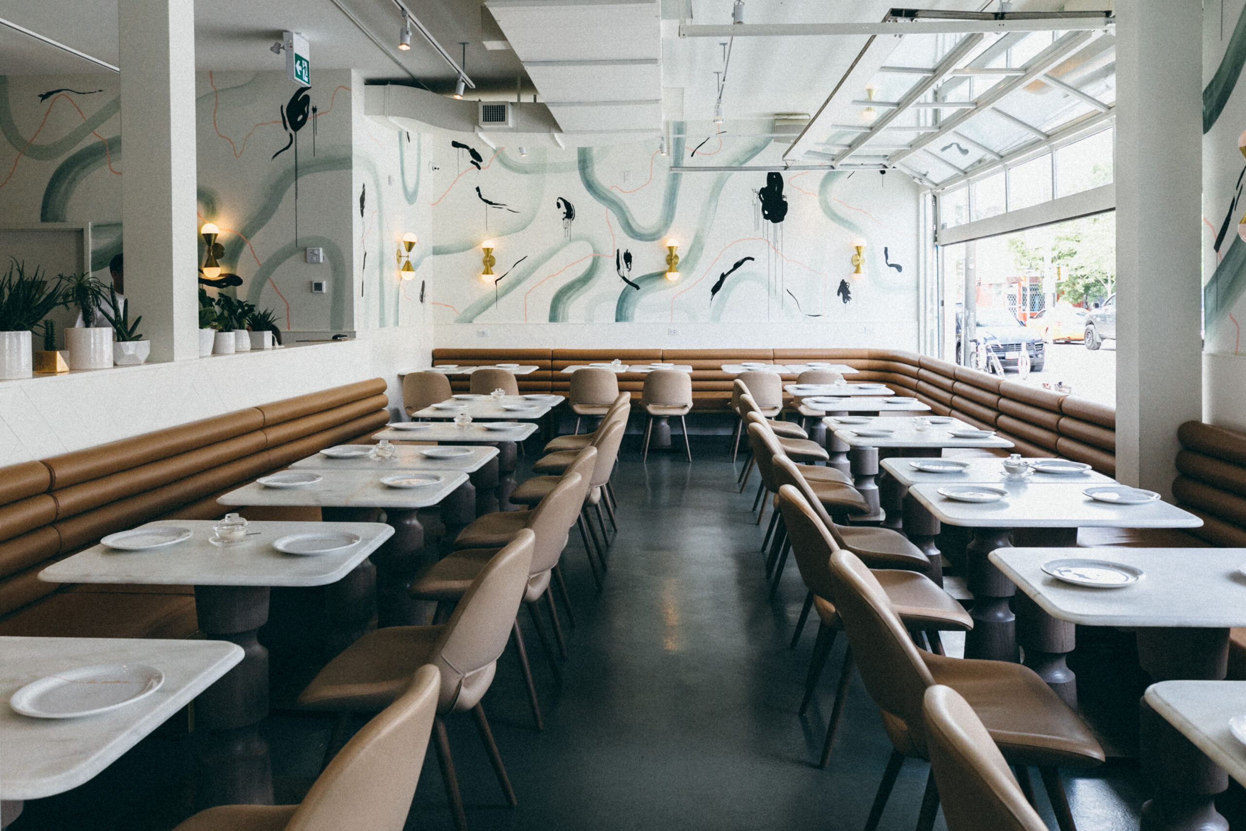 La Palma La Palma Toronto Restaurant Interior. White wall with pink and mint green streaks. White marble tables line the left, right, and back wall. There are caramel brown banquette backings behind the marble tables