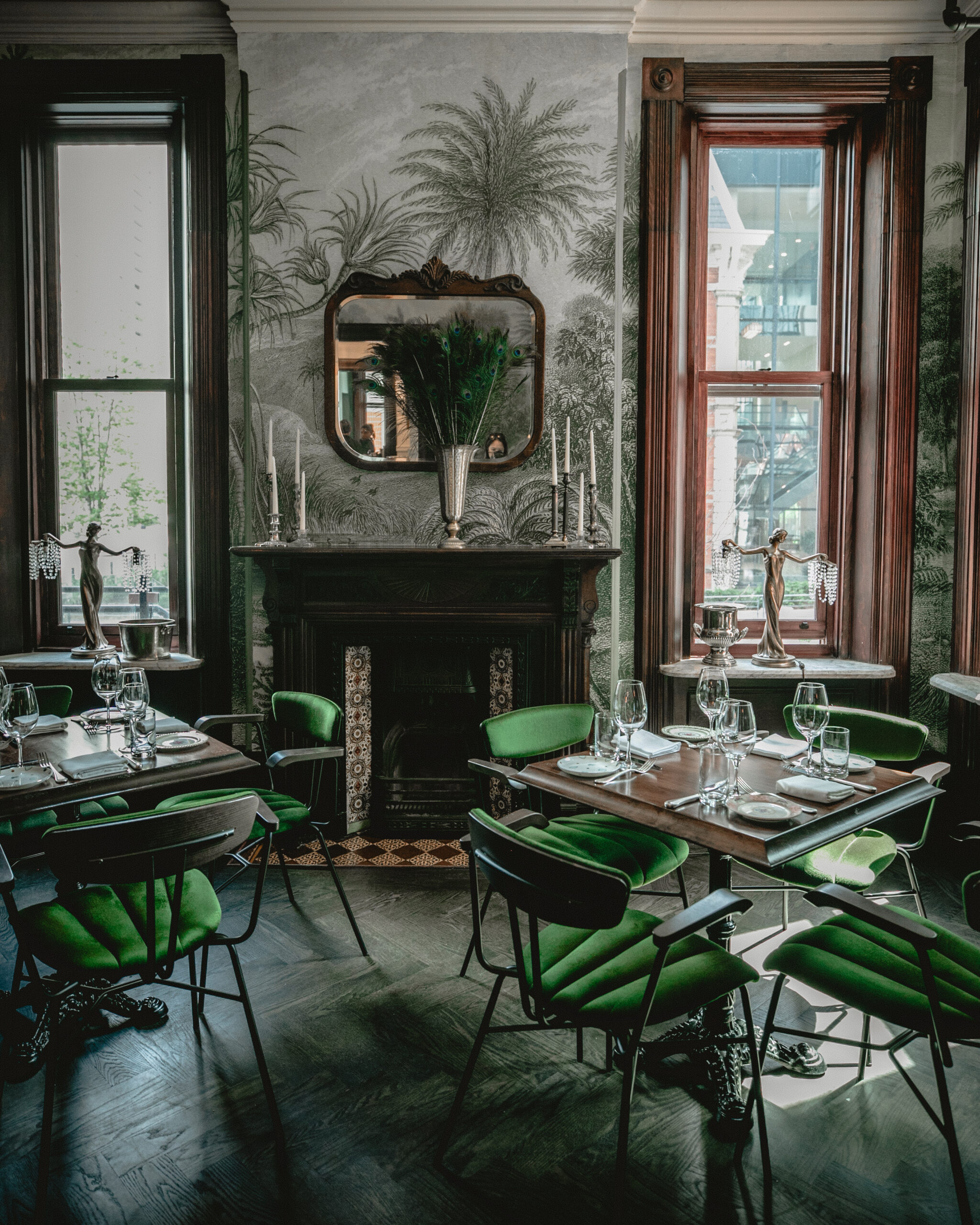 Maison Selby Main Room, velet green chairs surround wooden tables in a historic building. A decorative fireplace wooden mantle is on the back wall with vintage style wallpaper with thin brush stroke drawings of trees.A wooden frame square mirror hangs above the fireplace with two long rectangular windows on both sides.