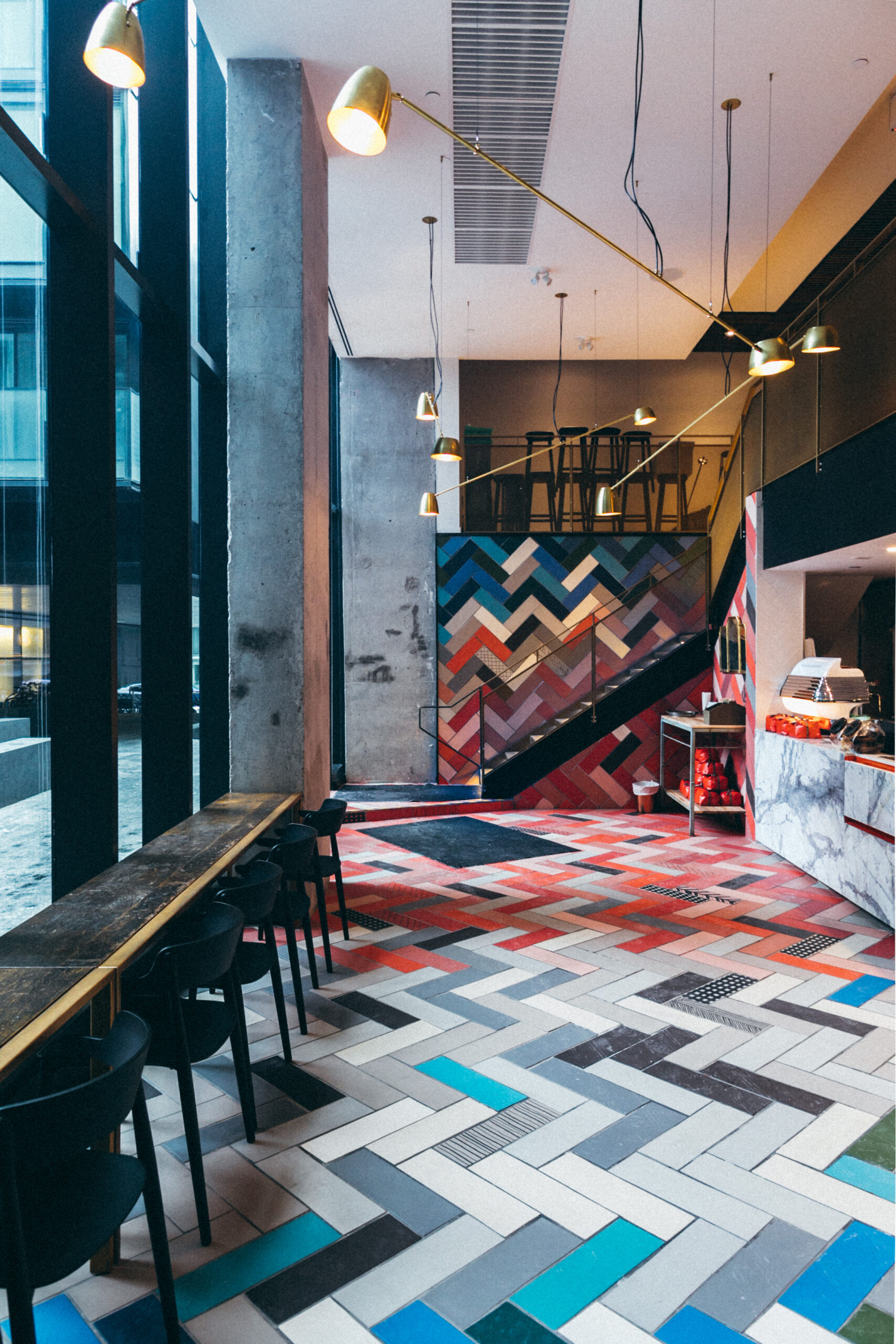 Oretta Restaurant Coffee Shop Toronto Interior. Blue, white, red, pink, long rectangular tiles laid out in herringbone patter on the floor. Wooden Bar seating lines the floor to ceiling windows. Coffee bar is on the right with staircase on the back wall that is also lined with same blue, white, red, pink tiles on the back wall