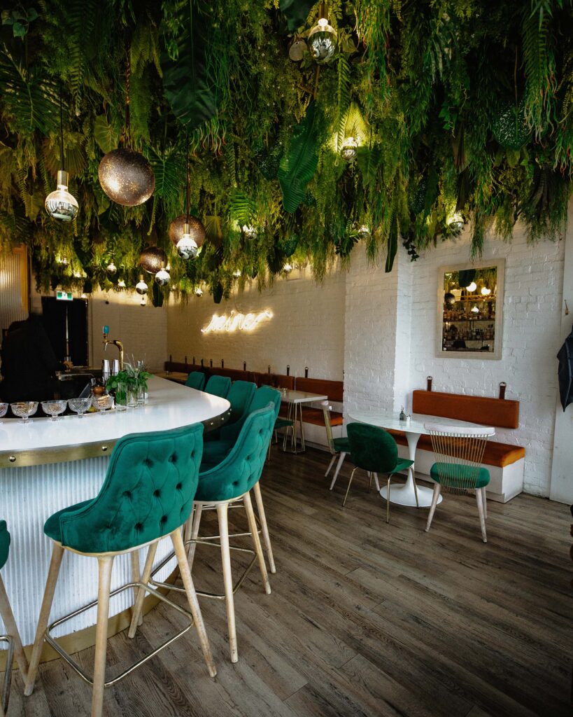 Reyna on King Beautiful Restaurant in Toronto. Emerald green velvet chairs with wooden legs line the white marble bar top. Red banquette seating is on white brick back wall with white marble tables. Greenery hangs from ceilings. There is a yellow neon sign on the back wall with the word "reine" written in cursive writing